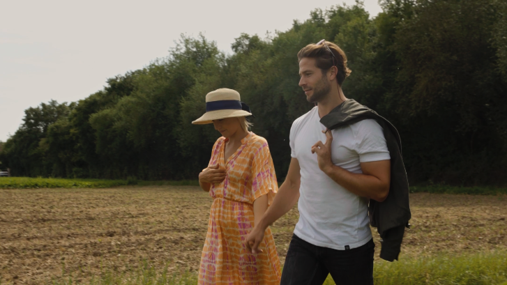 Paar hält sich an den Händen beim Spaziergang durch eine Wiese, sie im Sommerkleid mit Hut, er mit Rucksack.