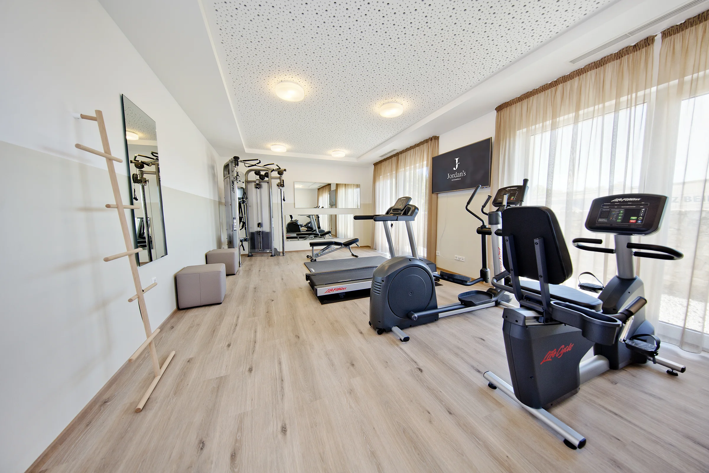 Heller Fitness- und Therapieraum mit mehreren Trainingsgeräten, großen Fenstern und hellem Holzboden.
